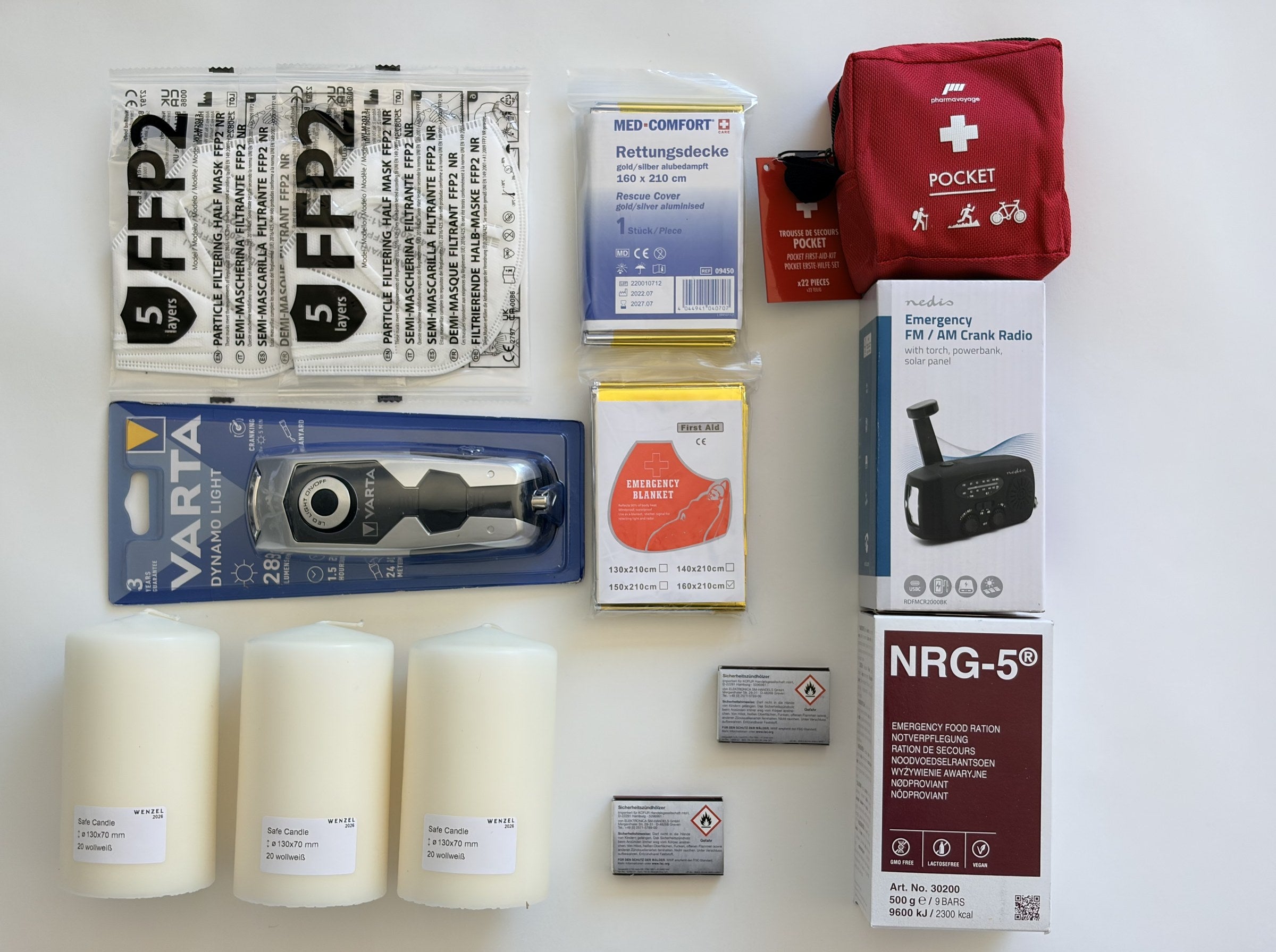 Der Inhalt der NV-Essentials im Raster ausgelegt: Kurbelradio, Kurbellampe, Notration, Erste-Hilfe-Set, drei Kerzen, zwei Rettungsdecken, zwei FFP2-Masken, zwei Schachteln Streichhölzer